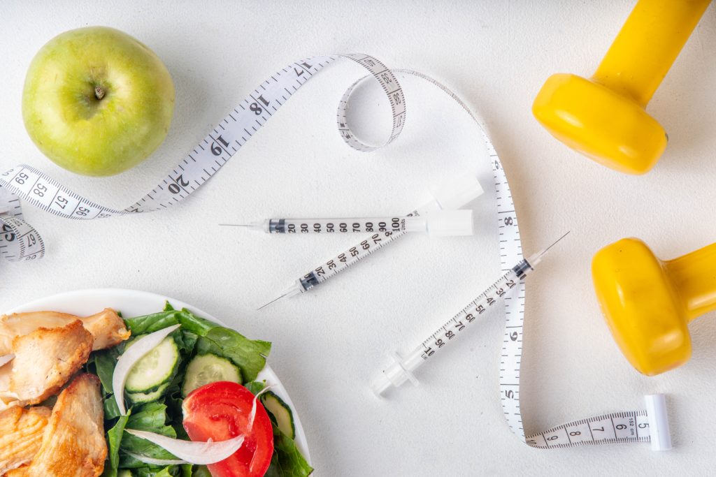 Seringas de medicação ao lado de fita métrica, halteres e alimentos saudáveis, representando o uso de tirzepatida e a relação com vitamina B12 no contexto de controle de peso e nutrição.