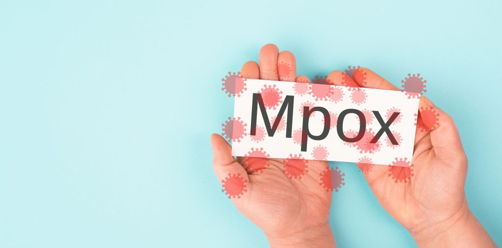 Pessoa segurando um cartão com a palavra “Mpox”, com ilustrações do vírus ao redor, sobre fundo azul.