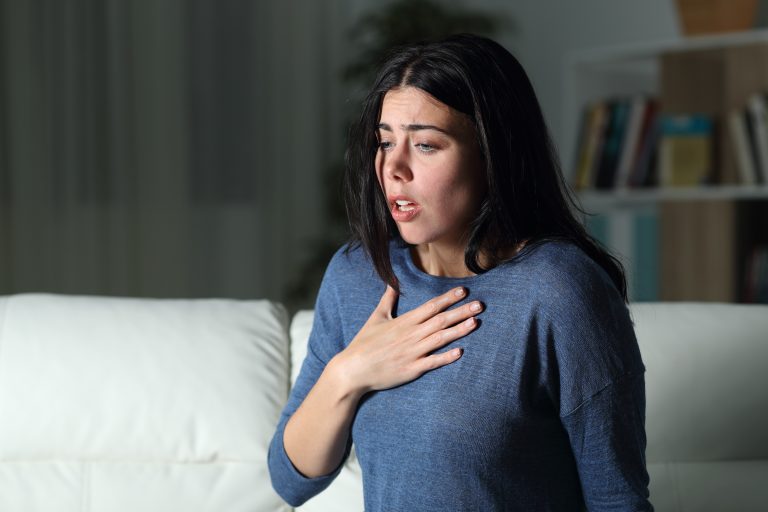 Mulher com expressão de preocupação e dificuldade para respirar, segurando o peito durante uma crise de ansiedade em ambiente doméstico.