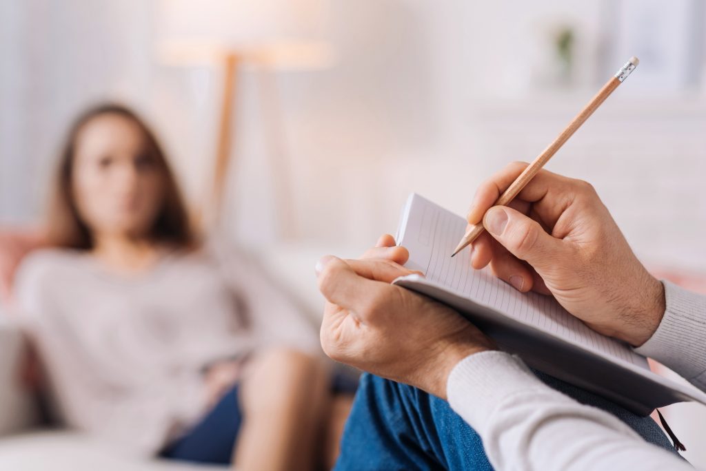 Profissional de saúde mental fazendo anotações durante sessão de terapia com paciente ao fundo, representando o acompanhamento de sintomas relacionados a borderline e bipolaridade.