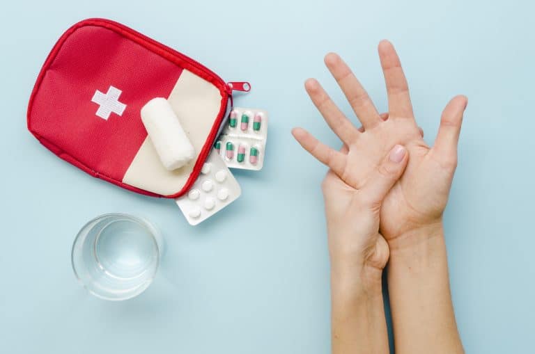 Kit de primeiros socorros com curativos, comprimidos e água ao lado de uma pessoa segurando a própria mão, ilustrando cuidados básicos em situações de emergência do dia a dia.