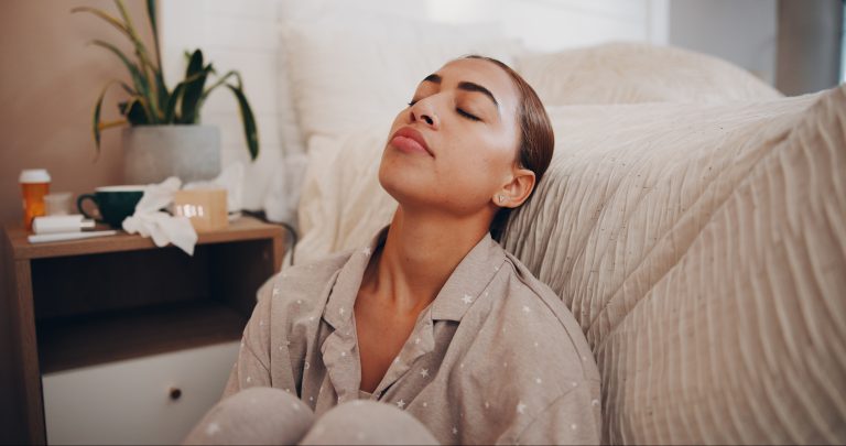 Pessoa sentada ao lado da cama com os olhos fechados e expressão de exaustão, representando o cansaço persistente que pode afetar a disposição diária e estar relacionado a diferentes condições de saúde física ou emocional.