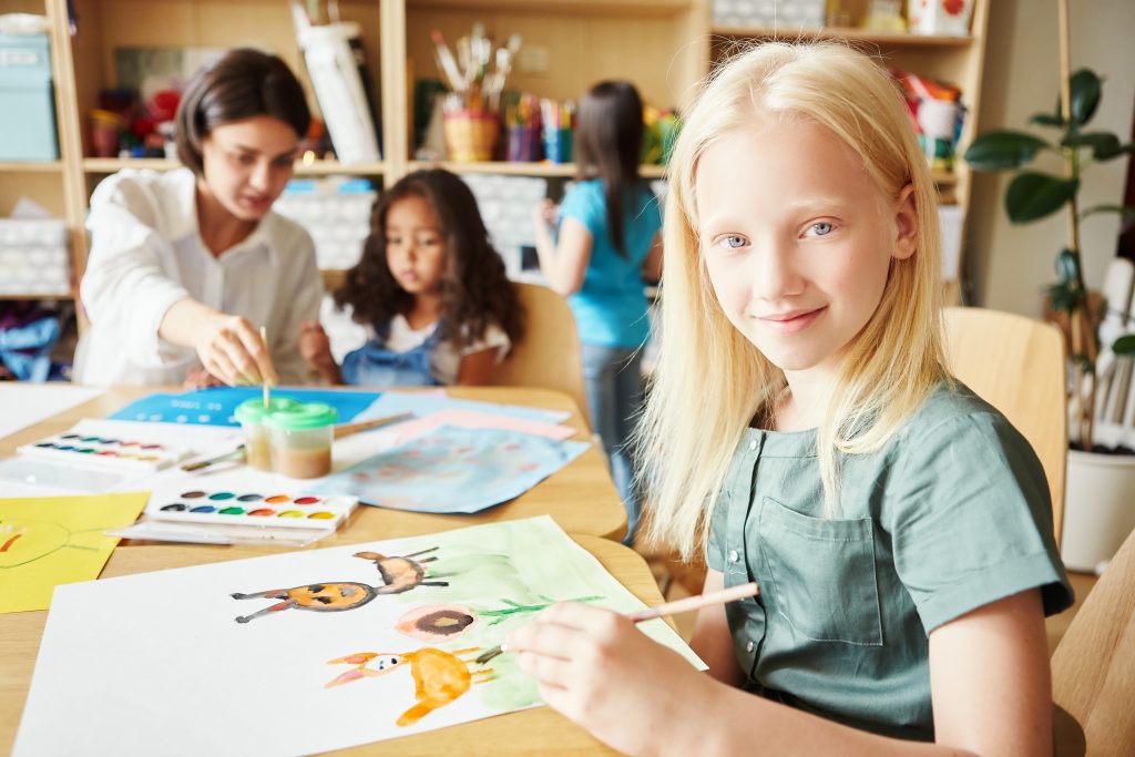 Criança com albinismo em sala de aula, pintando um desenho enquanto interage com colegas e educadora, destacando a importância da inclusão.