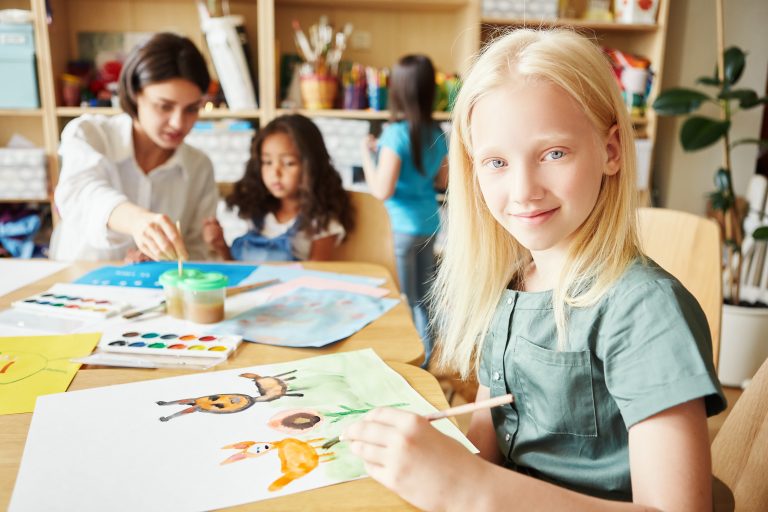 Criança com albinismo em sala de aula, pintando um desenho enquanto interage com colegas e educadora, destacando a importância da inclusão.