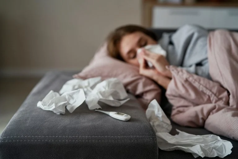 Pessoa com sintomas de gripe K deitada no sofá assoando o nariz, com termômetro e lenços usados ao lado.