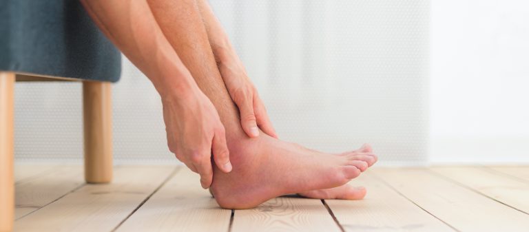 Pessoa sentada segura o tornozelo com as mãos, evidenciando inchaço na região do pé e da articulação, em ambiente interno com piso de madeira claro.