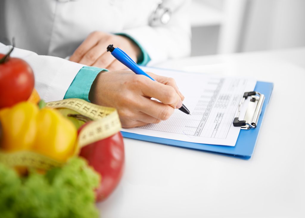Nutricionista registrando informações em um plano alimentar durante consulta, com frutas, vegetais e fita métrica em destaque, representando o acompanhamento nutricional para pessoas com intolerância alimentar e a busca por mais qualidade de vida.