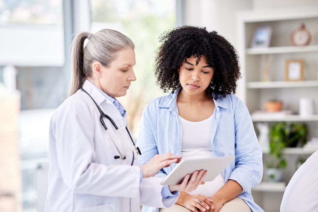 SOMP: o que é a Síndrome Ovariana Metabólica Poliendócrina e quais são os sintomas? 5 Médica apresenta informações em um tablet durante consulta com paciente, ilustrando a investigação diagnóstica e o acompanhamento dos sintomas relacionados à SOMP (Síndrome Ovariana Metabólica Poliendócrina).