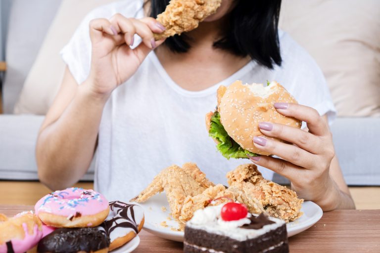 Pessoa consumindo hambúrguer, frango frito e doces, ilustrando o comer emocional, quando a alimentação é usada para aliviar emoções em vez de saciar a fome.