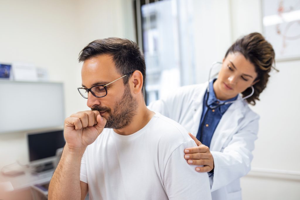 Pessoa com tosse sendo examinada por uma médica com estetoscópio, ilustrando a avaliação clínica para diagnóstico da pneumonia diante de sintomas respiratórios persistentes.