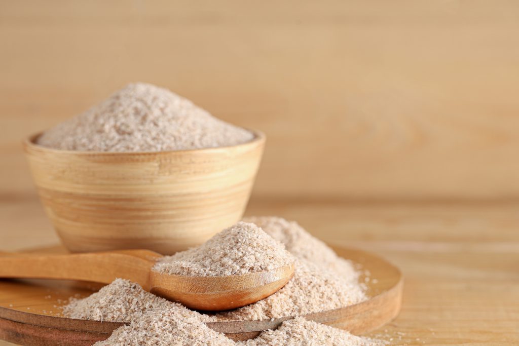 Psyllium em pó disposto em colher e recipiente de madeira sobre superfície clara, ilustrando o suplemento rico em fibras frequentemente utilizado para promover saciedade e auxiliar o funcionamento intestina.