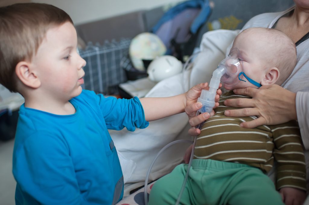 Bebê recebendo nebulização com máscara enquanto está no colo de um adulto, acompanhado por outra criança, ilustrando o cuidado com a bronquiolite e outras infecções respiratórias na infância.